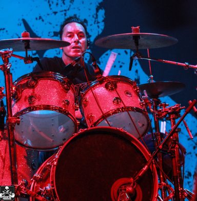 A drummer playing on a set against a vibrant blue and red backdrop.