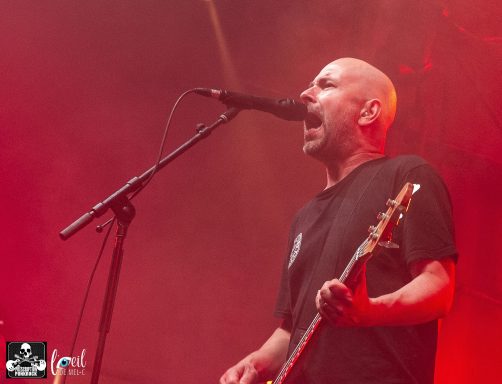 A male musician sings passionately into a microphone on stage with red lighting.