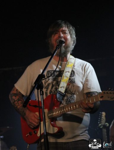 A man with tattoos sings and plays a red guitar on stage.
