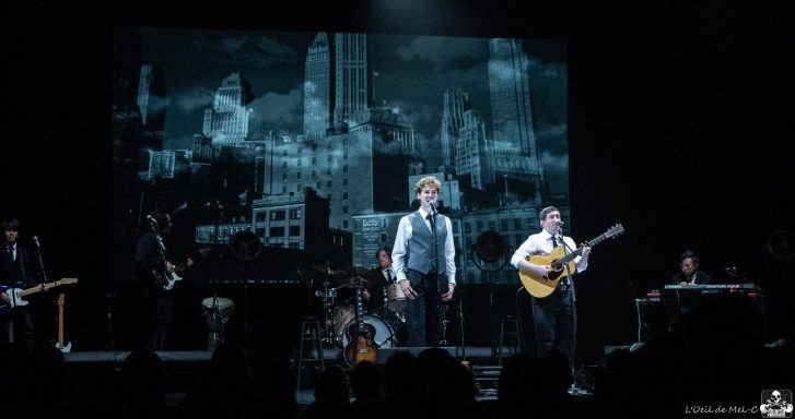 Two performers on stage with instruments, backdrop features a cityscape.