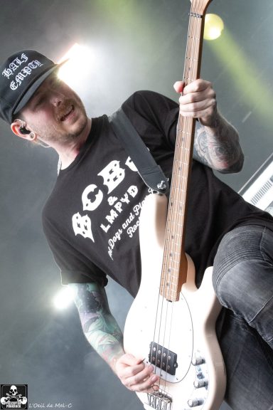 Musician playing a white bass guitar on stage, wearing a black cap and shirt.