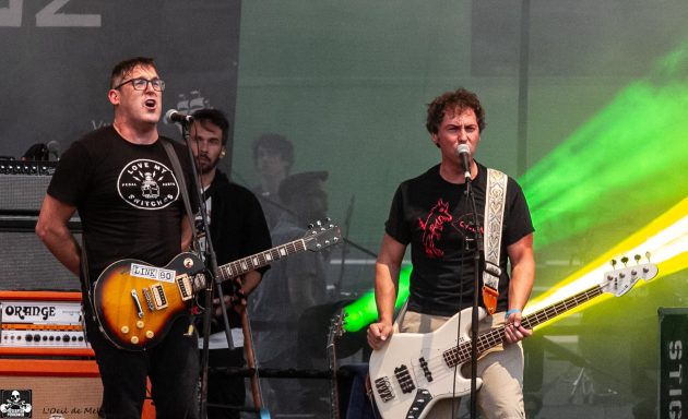 Two musicians perform on stage with electric guitars and vibrant lights in the background.