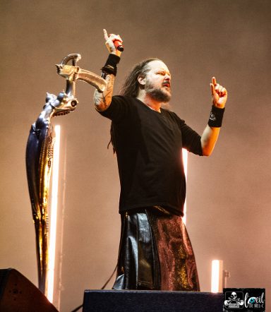 A musician wearing a black shirt and kilt performs on stage, raising one hand.