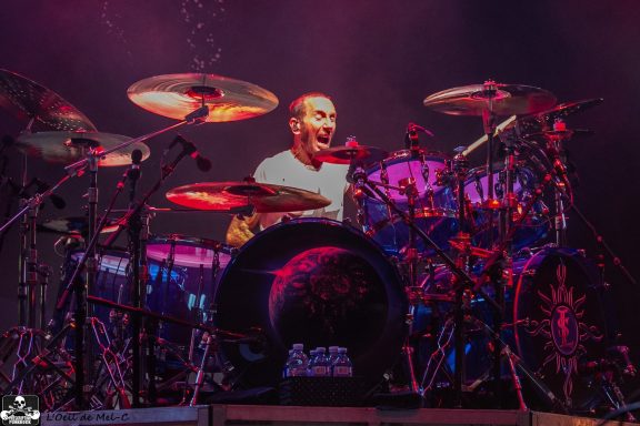 Drummer performing energetically on stage with colorful lighting.