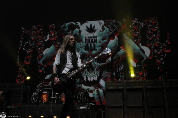 A bassist performs on stage in front of a dramatic backdrop featuring a skull and vibrant graphics.