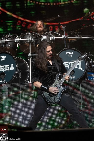 Guitarist performing energetically on stage with a drummer in the background.