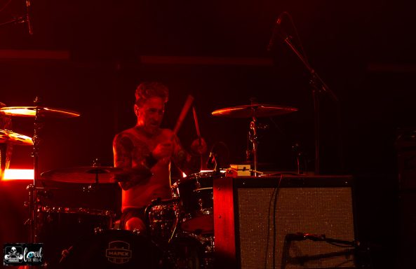 A drummer performing on stage under red lighting.