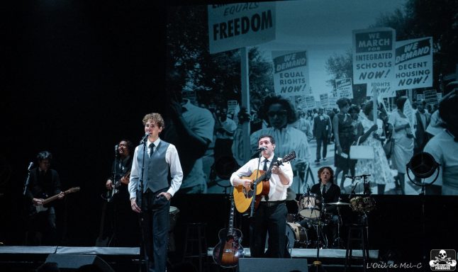 Musicians performing on stage with a backdrop of a historical civil rights protest.
