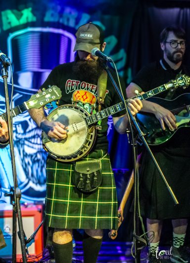 Man in a kilt playing a banjo with musicians in the background.