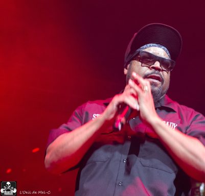 A performer with glasses and a bandana holds a microphone in a vibrant red-lit setting.