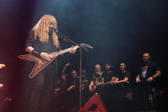Musician playing electric guitar on stage with an enthusiastic crowd in the background.
