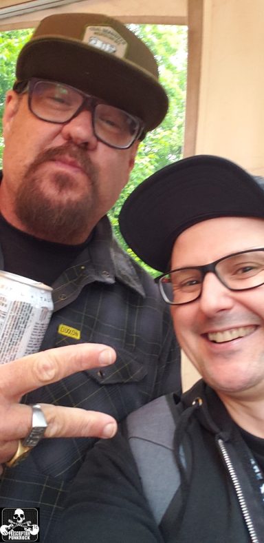 Two men posing together, one holding a can, both wearing hats and glasses.