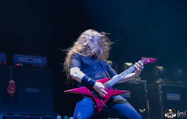 Musician with long hair playing a red electric guitar on stage.
