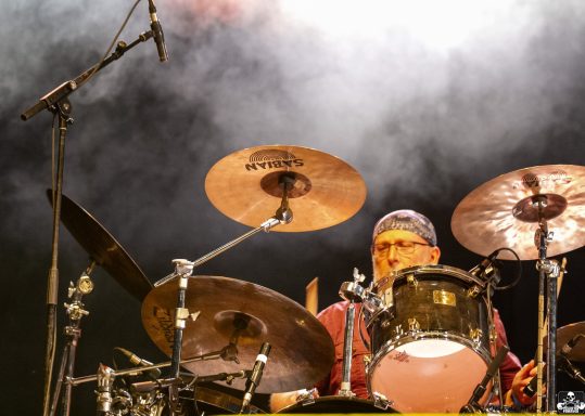 Drummer performing on stage with cymbals and dramatic lighting.
