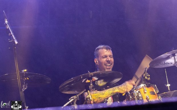 Drummer performing energetically on stage with a vibrant purple background.