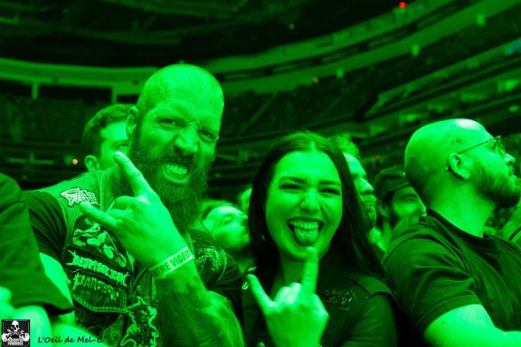 Two people making rockhand gestures at a concert, surrounded by fans in a vibrant green light.