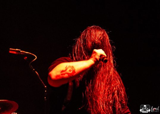 A musician with long hair performs passionately against a dark background.