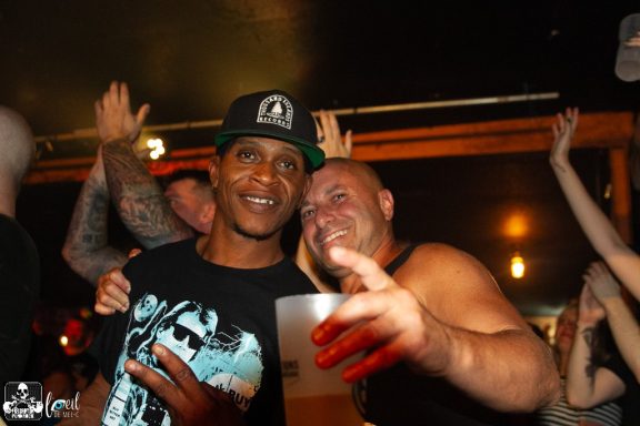Two men smiling and posing together in a lively bar setting, with people celebrating in the background.