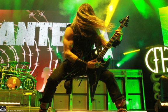 Guitarist performing energetically on stage with bright lights in the background.