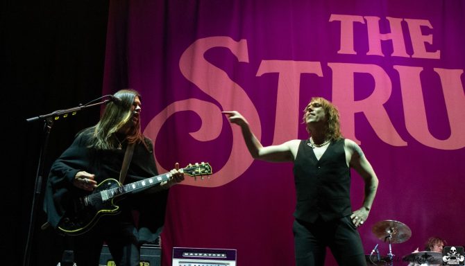 Two musicians perform on stage, with a prominent backdrop featuring "The Struts."