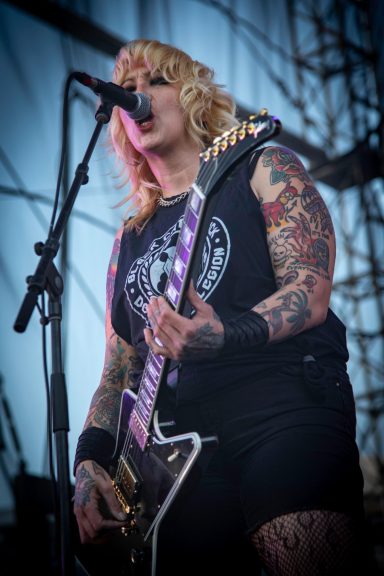 Female guitarist with tattoos performs on stage, wearing a black tank top and shorts.