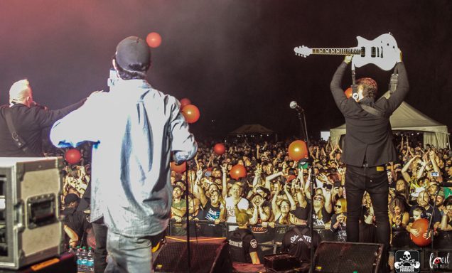 Two musicians on stage face a large crowd with balloons at an outdoor concert.