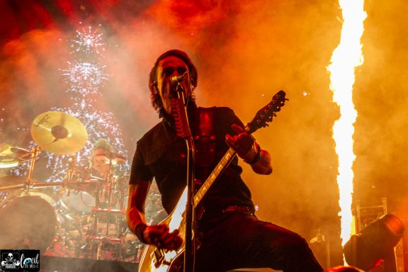A guitarist performing on stage with dramatic lighting and pyrotechnics in the background.