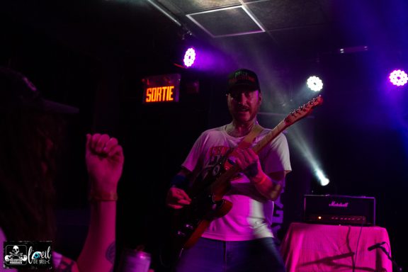 Musician playing electric guitar on stage with colorful lighting and audience hand raised.