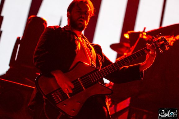 Musician playing a guitar on stage, illuminated by red lights.