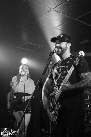 A musician with a guitar performs on stage while a vocalist sings nearby, both smiling.