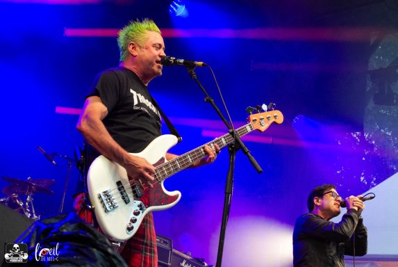 A musician with green hair plays bass while another sings on stage.
