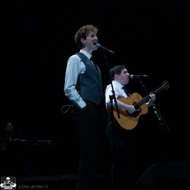 Two performers on stage singing, one playing guitar, with a dark background.