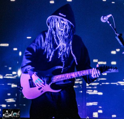 A musician in a dark hoodie plays an electric guitar against a blue-lit backdrop.