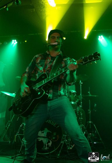 Musician performing on stage with a guitar, under green and yellow stage lights.
