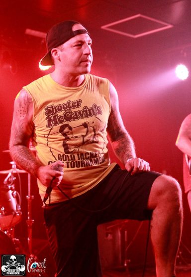 Musician performing on stage in a yellow tank top, with microphone in hand.