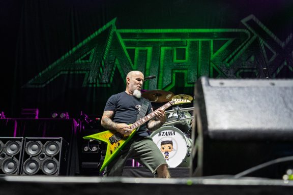 Musician playing guitar on stage with band logo in the background.