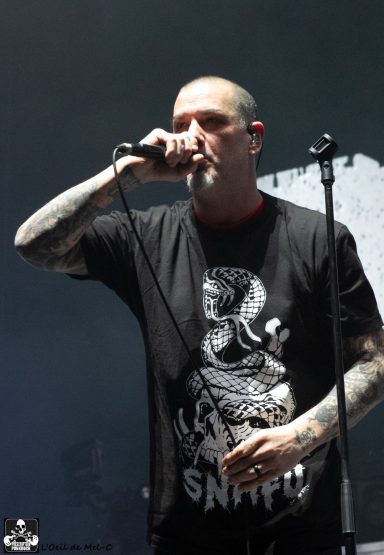 Man with tattoos performs on stage, holding a microphone and wearing a graphic t-shirt.