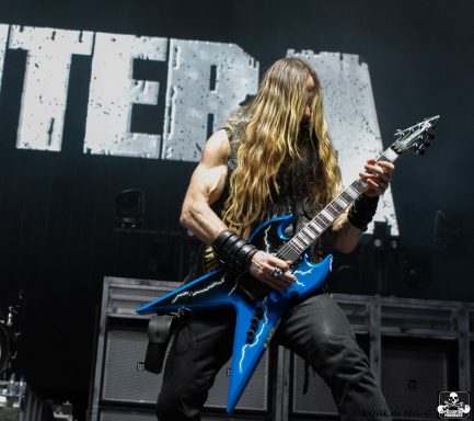Musician playing a blue electric guitar with long hair on stage in front of a backdrop.