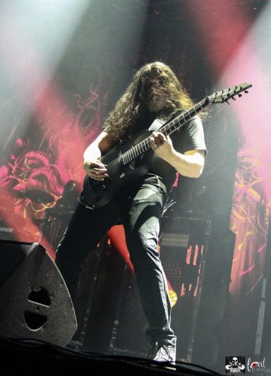 Musician with long hair playing electric guitar on stage with dramatic lighting.
