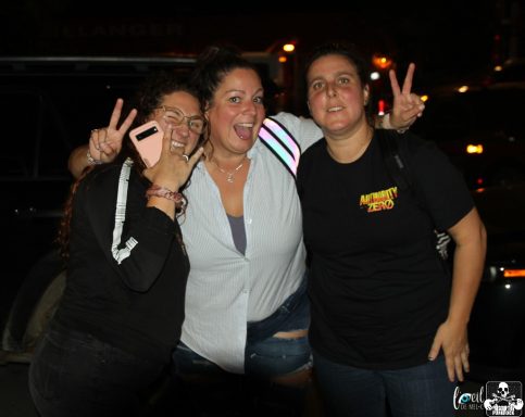 Three women posing together, smiling and showing peace signs outdoors at night.