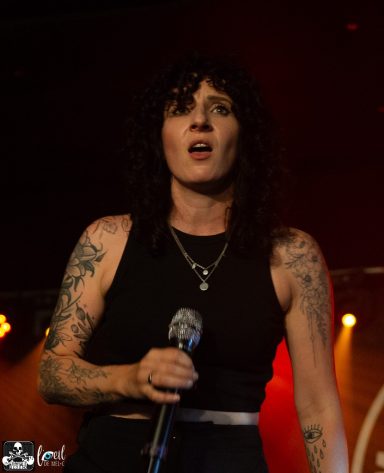 A woman with curly hair and tattoos sings into a microphone on stage.