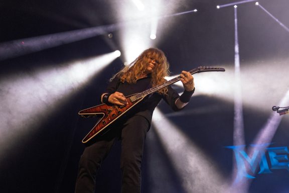 Musician playing electric guitar under stage lights, with long hair and intense focus.