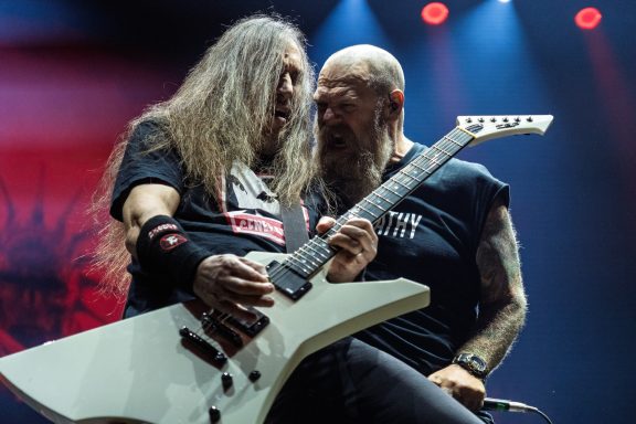 Two male musicians perform passionately on stage, one playing an electric guitar.