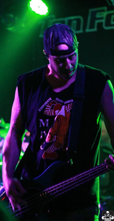 A musician playing bass guitar on stage, wearing a sleeveless shirt and cap.