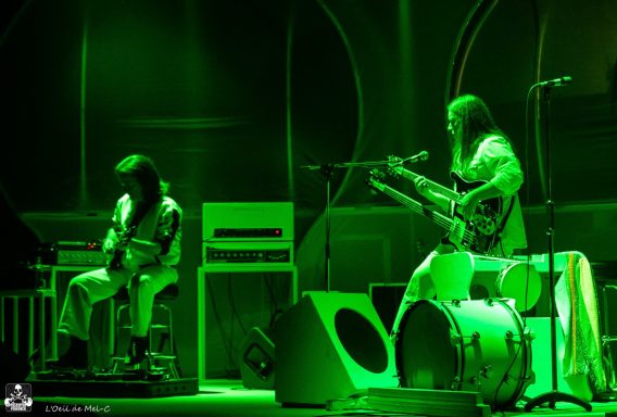 Two musicians perform on stage, illuminated by green lighting, with instruments around them.