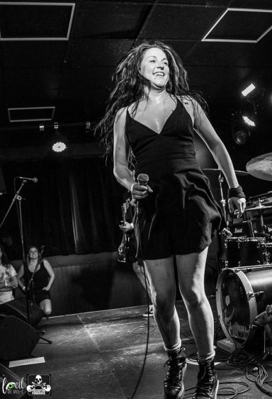 Woman in a black dress performs on stage with a microphone, surrounded by musicians.