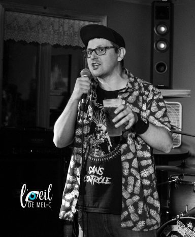 Man in a patterned shirt and cap, speaking into a microphone, holding a drink.