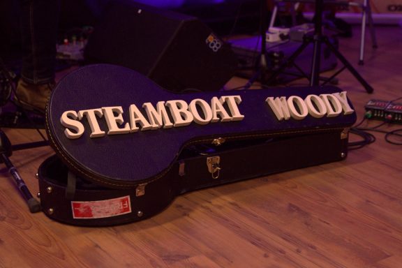 Black guitar case with "STEAMBOAT WOODY" written on it, resting on a wooden floor.