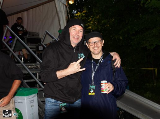 Two men pose together, one making a hand gesture, with a drink in hand.