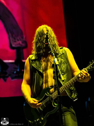 A musician with long hair in a sleeveless vest plays electric guitar on stage.